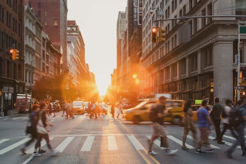 A bustling city street at sunset, with people crossing and yellow taxis passing by. Sunlight filters between tall buildings, creating a warm, vibrant glow
