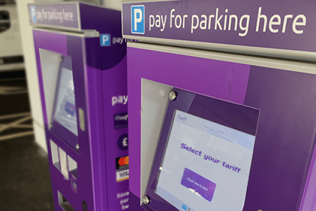 Purple parking payment kiosks with digital screens prompting "Select your tariff." Signs above read "pay for parking here," indicating ease and accessibility.