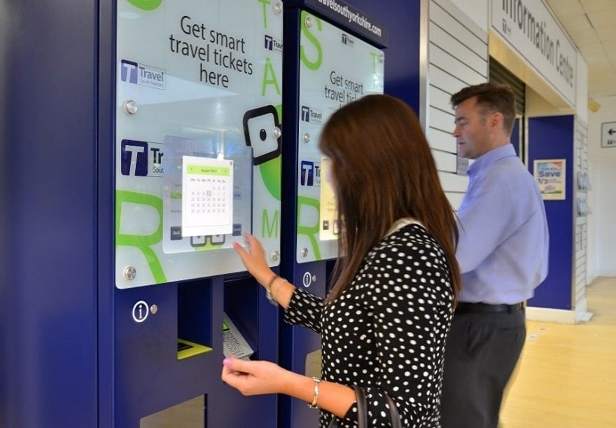 A woman in a polka dot dress and a man use sleek, modern ticket machines in a bright travel center, conveying convenience and efficiency.