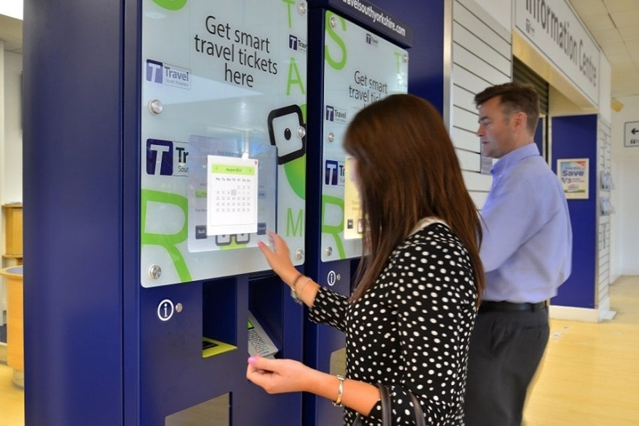 A woman in a polka dot dress and a man use sleek, modern ticket machines in a bright travel center, conveying convenience and efficiency.