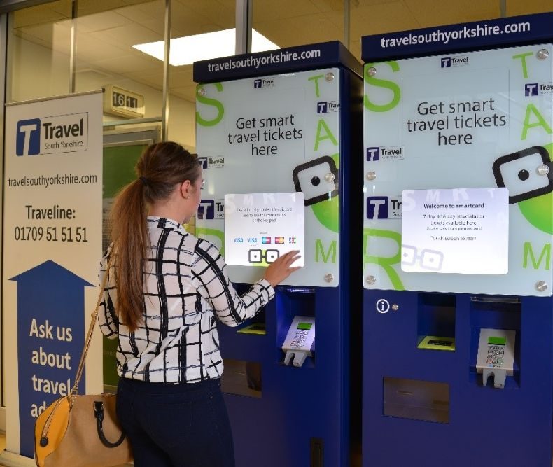 A woman interacts with a blue ticket machine at a travel office. The screen displays options for smart travel tickets. The setting is modern and organized.