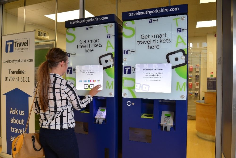 A woman interacts with a blue ticket machine at a travel office. The screen displays options for smart travel tickets. The setting is modern and organized.