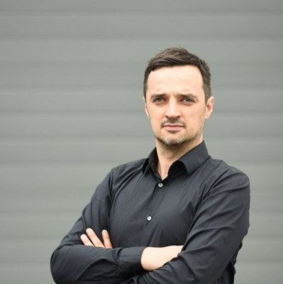 A man in a black shirt stands confidently with arms crossed against a gray backdrop. His expression is serious, conveying professionalism and focus.