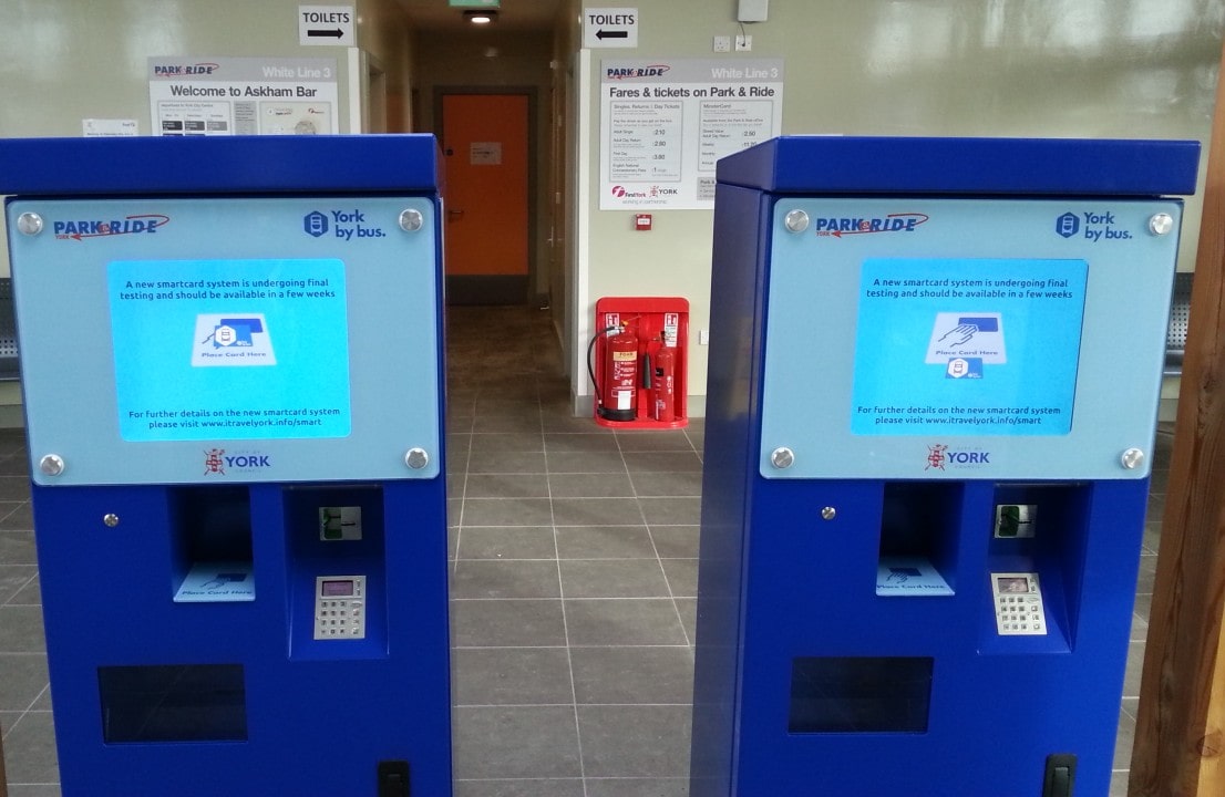 Two blue Park & Ride ticket machines with digital screens stand in a waiting area. Signs on the walls provide fare and service information.