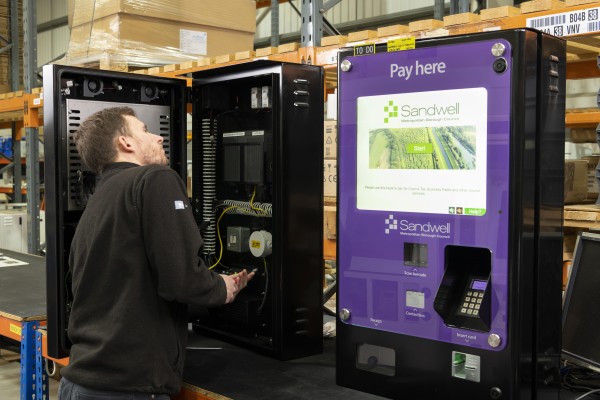 A technician inspects an open parking payment machine in a warehouse. The machine displays "Pay here" with a purple and white design. Shelves and boxes are stacked in the background.