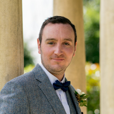 A man in a grey suit with a blue bow tie stands confidently, smiling softly. Sunlit greenery and columns in the background convey a warm, formal setting