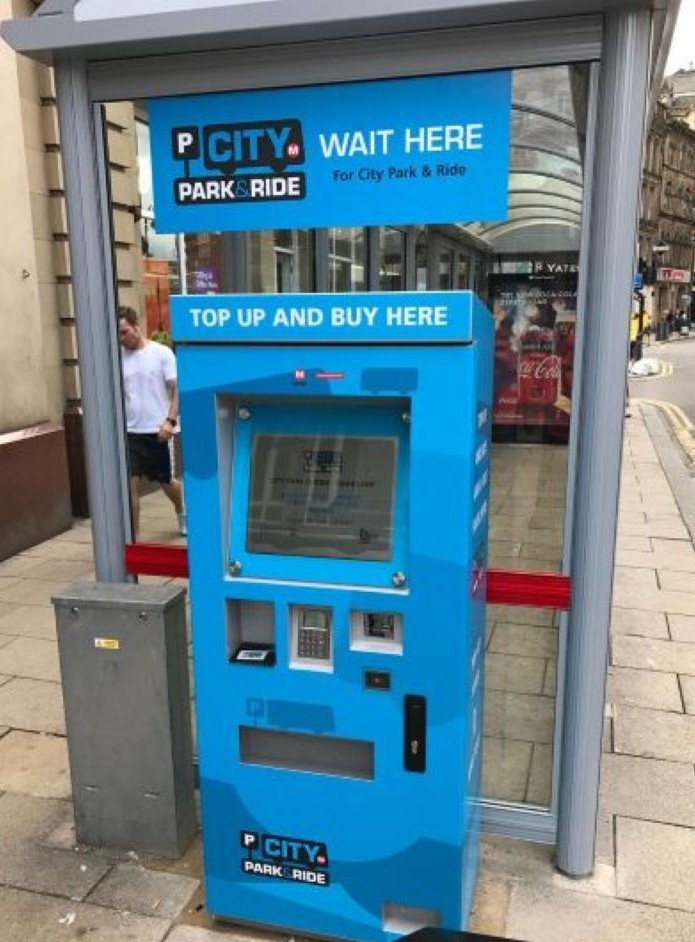 A blue "City Park & Ride" ticket machine stands at a bus stop with "Top Up And Buy Here" signage. Nearby, a pedestrian walks on a city street.
