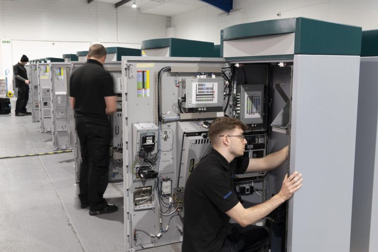 Technicians in a workshop work on tall electrical panels, focused and attentive. The room is well-lit, with a row of similar panels.