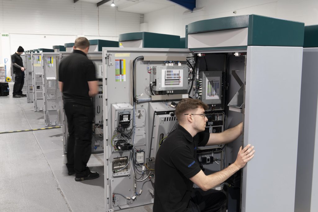 Technicians in a workshop work on tall electrical panels, focused and attentive. The room is well-lit, with a row of similar panels.