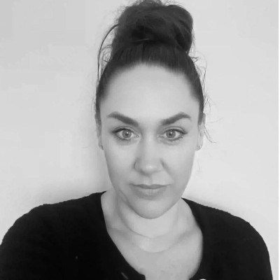 A woman with dark hair in a bun, wearing a dark top, looks directly at the camera with a neutral expression. The background is plain and light-colored.