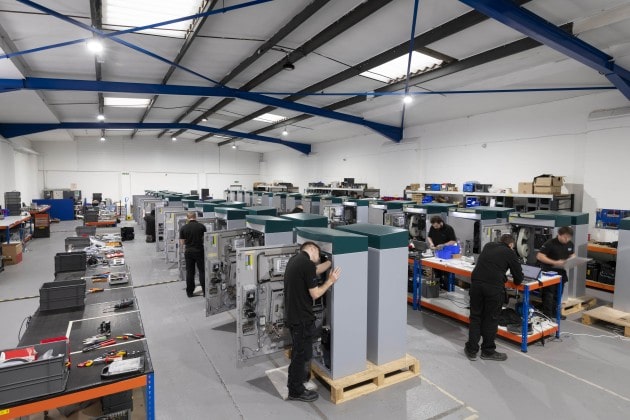 Workers in a spacious factory assemble large, grey machines. Bright lighting and organized rows create a focused and productive atmosphere.
