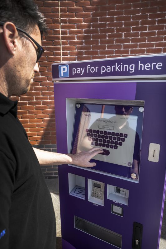 A man in sunglasses uses a modern, purple parking payment kiosk with a touchscreen under a sign that reads "pay for parking here." The setting is outdoors, with a brick wall in the background.