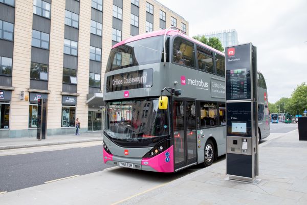Double-decker Metrobus with pink accents at a modern urban bus stop. Nearby, a digital timetable display stands. The scene is calm and bright.