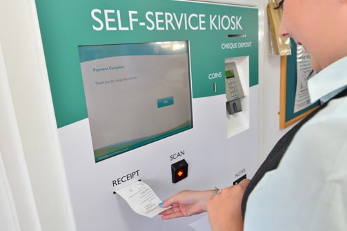 A person interacts with a self-service kiosk, retrieving a receipt. The screen displays a completed payment message. The mood is efficient and modern.