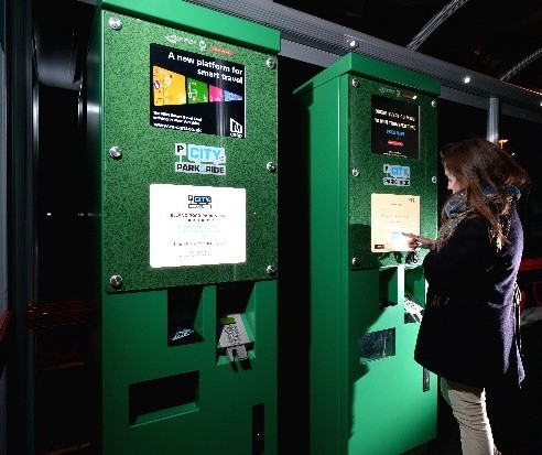 A woman uses a green parking ticket machine at night, pressing buttons on the touchscreen. The scene is well-lit, suggesting a modern, urban setting.