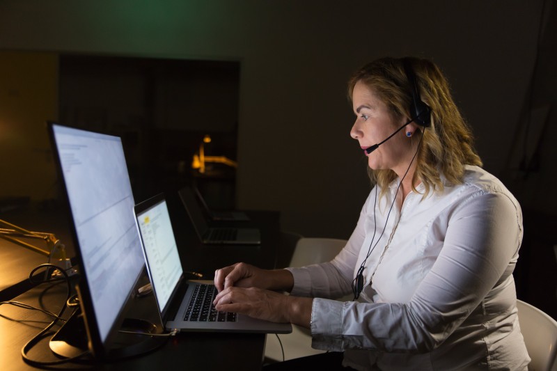 A woman in a white shirt works at a desk with two monitors and a laptop, wearing a headset. The setting is dimly lit, suggesting a focused, late-night work environment.