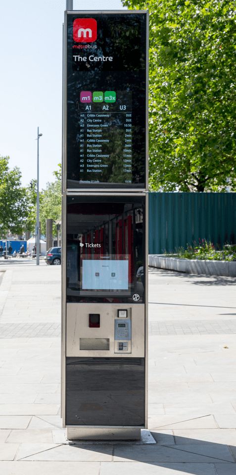 A modern bus ticket machine outdoors on a sunny day, displaying routes and times. Green trees and an urban street are visible in the background.